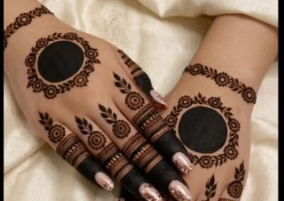 Two hands with circular mehndi designs on the back and detailed finger patterns, resting on a light fabric background.”