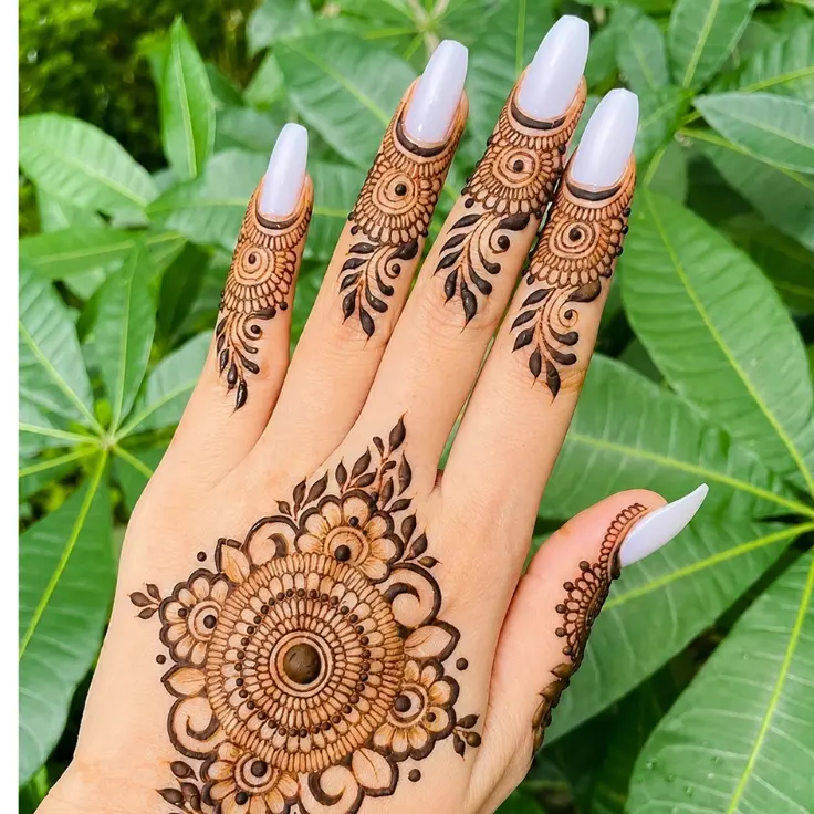 “Hand with circular floral mehndi design and decorated fingers over green leaves background.”