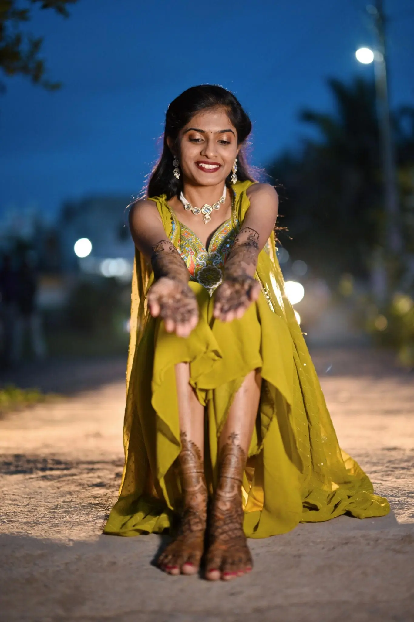 Young woman in a green traditional dress sitting on a road at dusk, smiling and extending her hands forward to display intricate mehndi designs on her palms and feet, with soft lights blurred in the background.